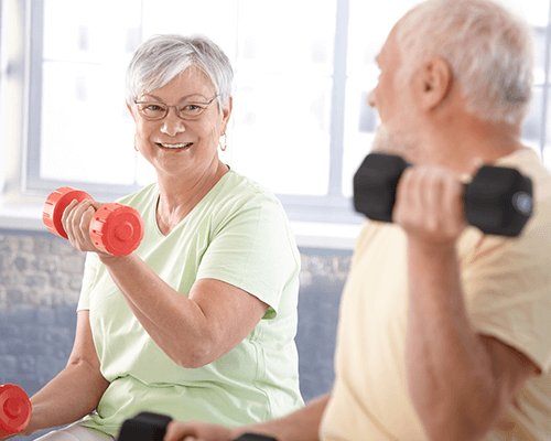 A happy elderly woman working out while smiling and looking at an elderly man also exercising close by in the foreground.